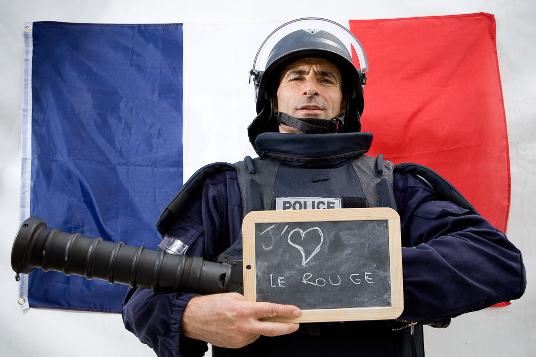 Police officer in riot gear poses for a photograph in front of the French flag holding a gun and a chalkboard with I Love Red Wine written there in French