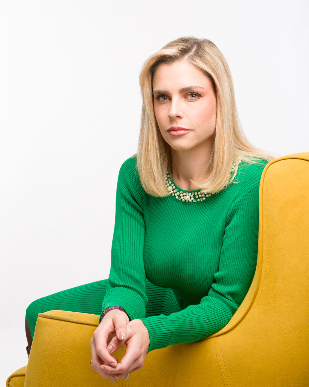 Studio photograph of blonde woman in a green dress sitting in a yellow armchair